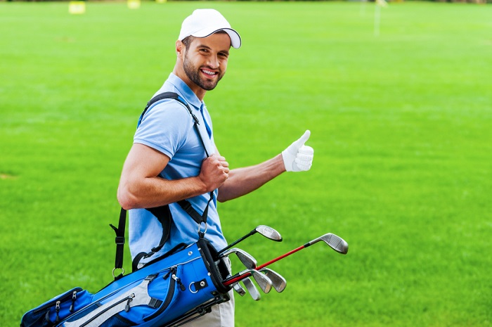 I love golfing! Rear view of young happy golfer carrying golf bag with drivers and looking over shoulder while standing on golf course